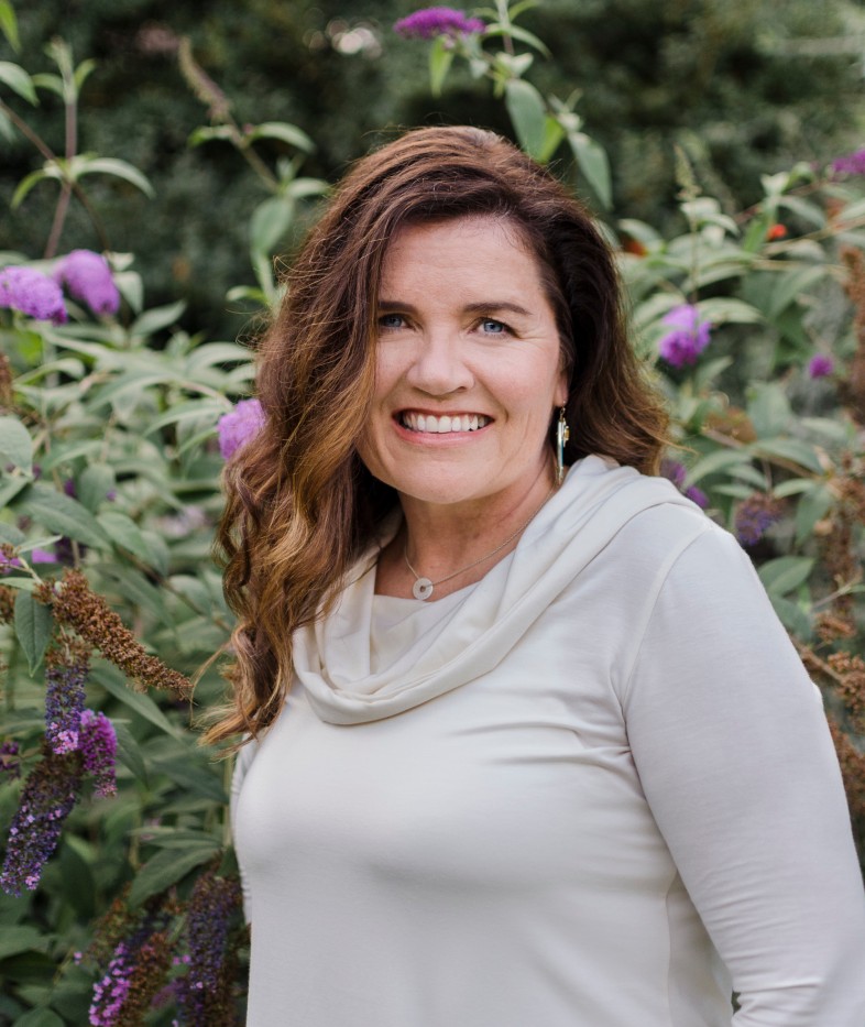 Woman stands against green shrub and purple flowers, smiling in headshot.