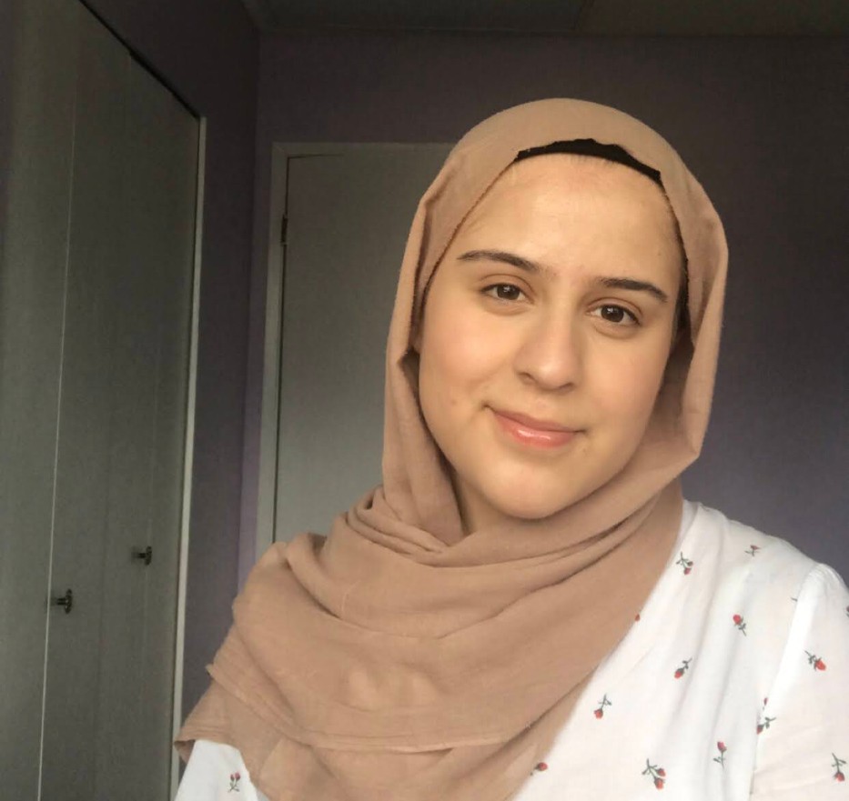 Selfie of woman in beige hijab and white patterned shirt, smiling.
