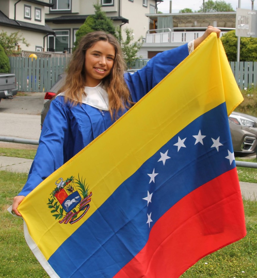 Woman standing in blue graduation gown, holding a large Venezuelan flag, smiling.