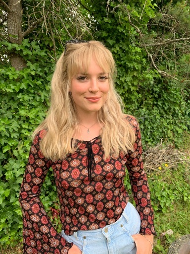 Blonde woman standing against green shubbery, smilng.