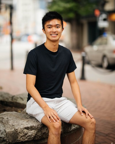 Young man in a black t-shirt is seated on a concrete ledge in the city, smiling.