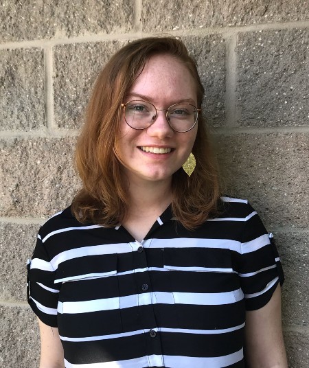 Young woman wearing black and white striped shirt stands against a brick wall, smiling,