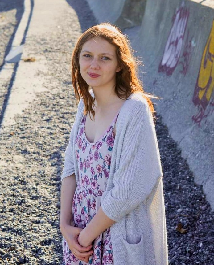 Young woman with brown hair stands in the sunlight, wearing a white cardigan with a floral patterned shirt.