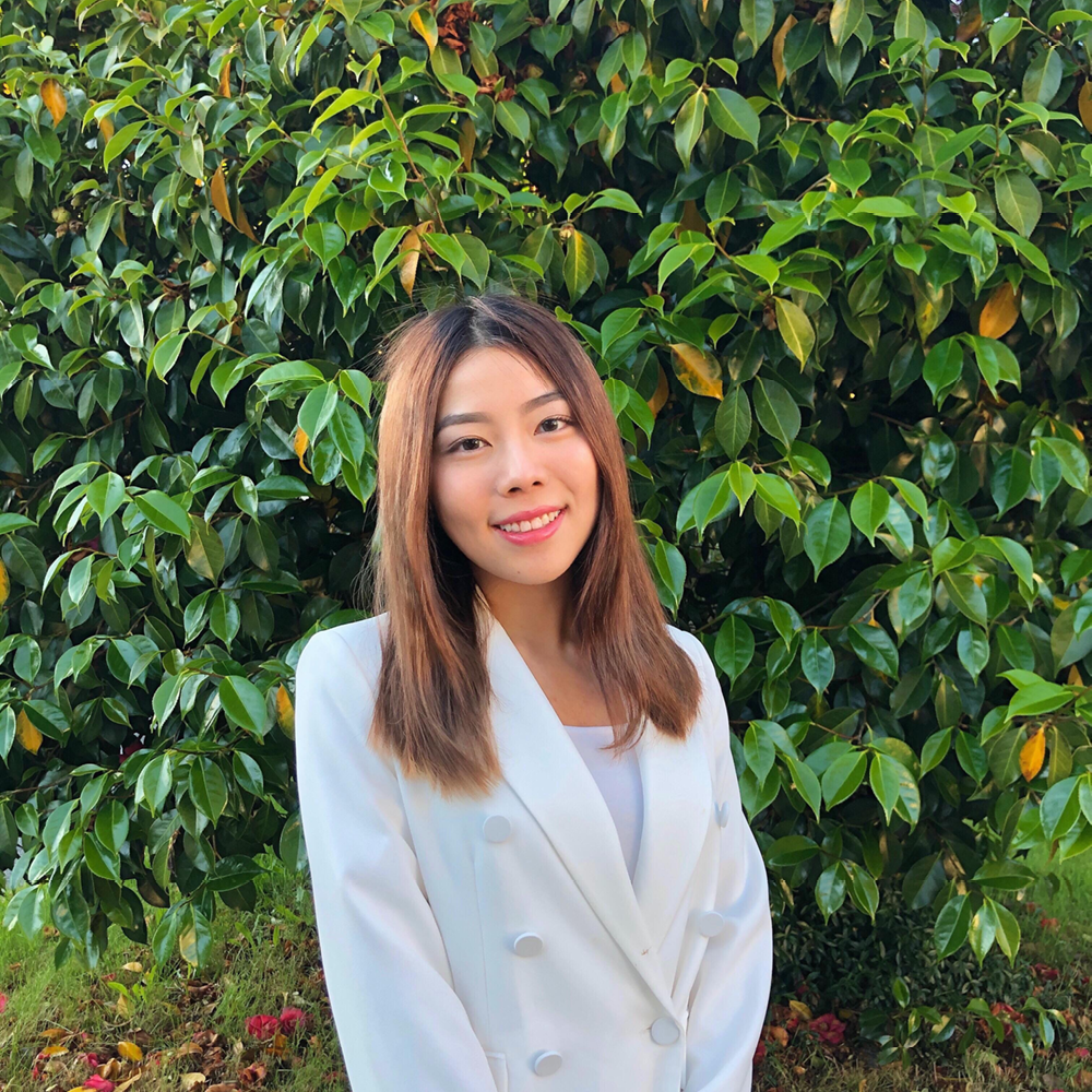 Young woman in white blazer smiles, standing against a background of greenery.