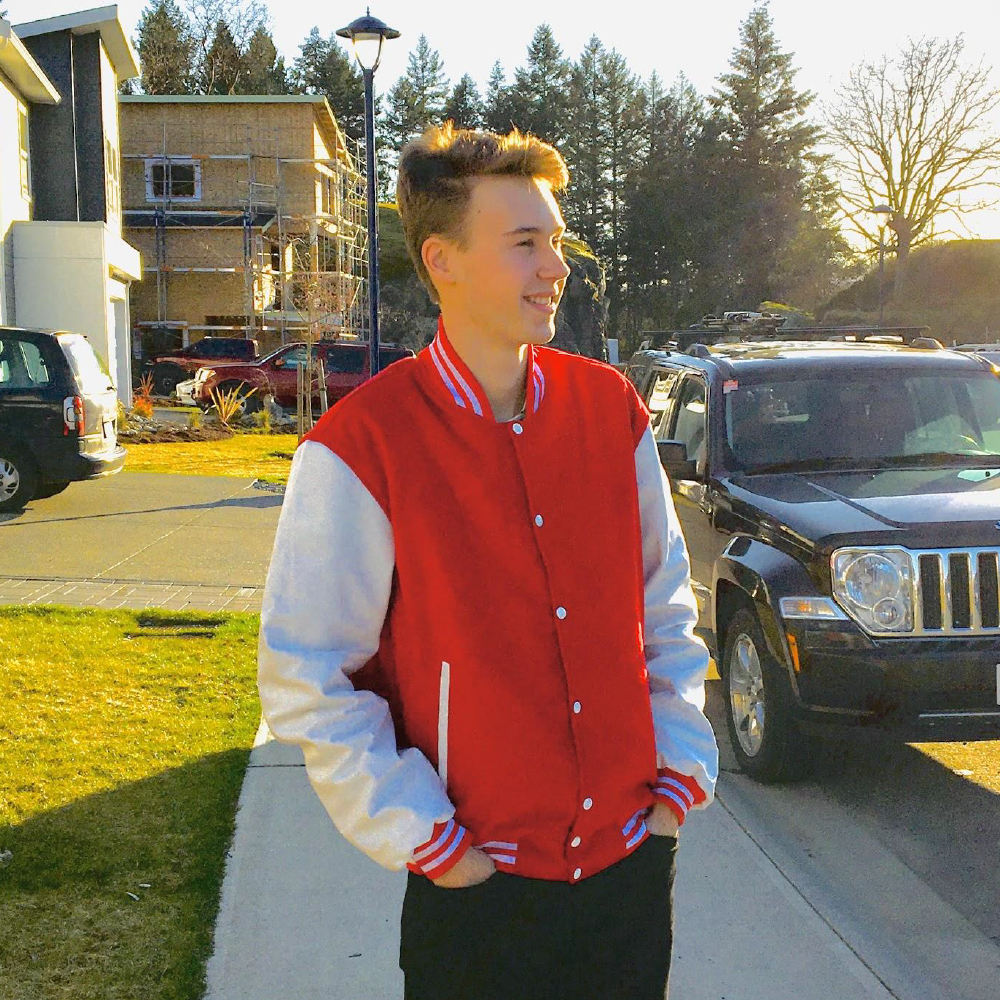 Young man wearing a red and white varisty jacket, stands in the middle of a neighbourhood sidewalk with two hands in his pockets while looking to the side.