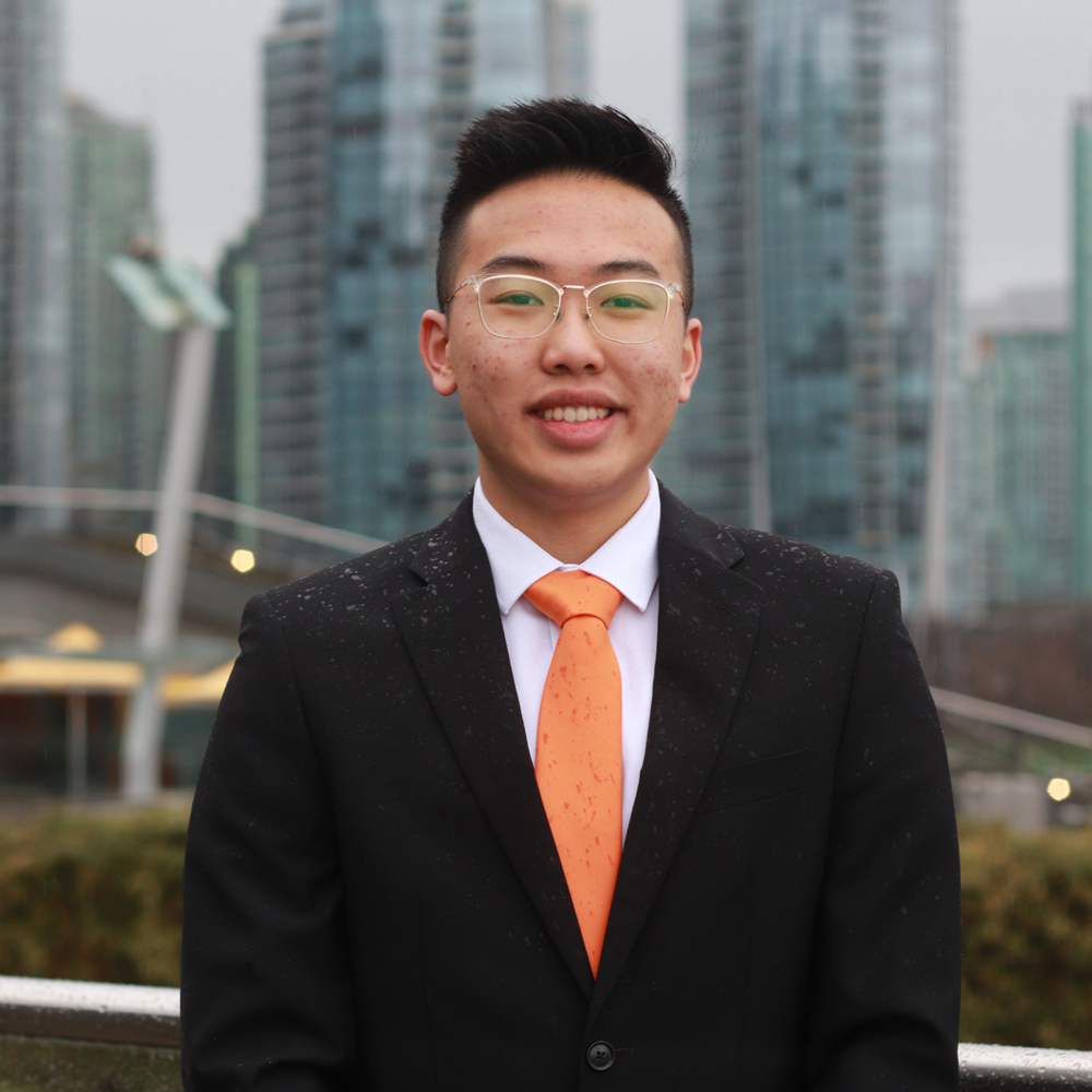Young man in a suit with orange tie smiles, standing in front of an array of highrise towers.