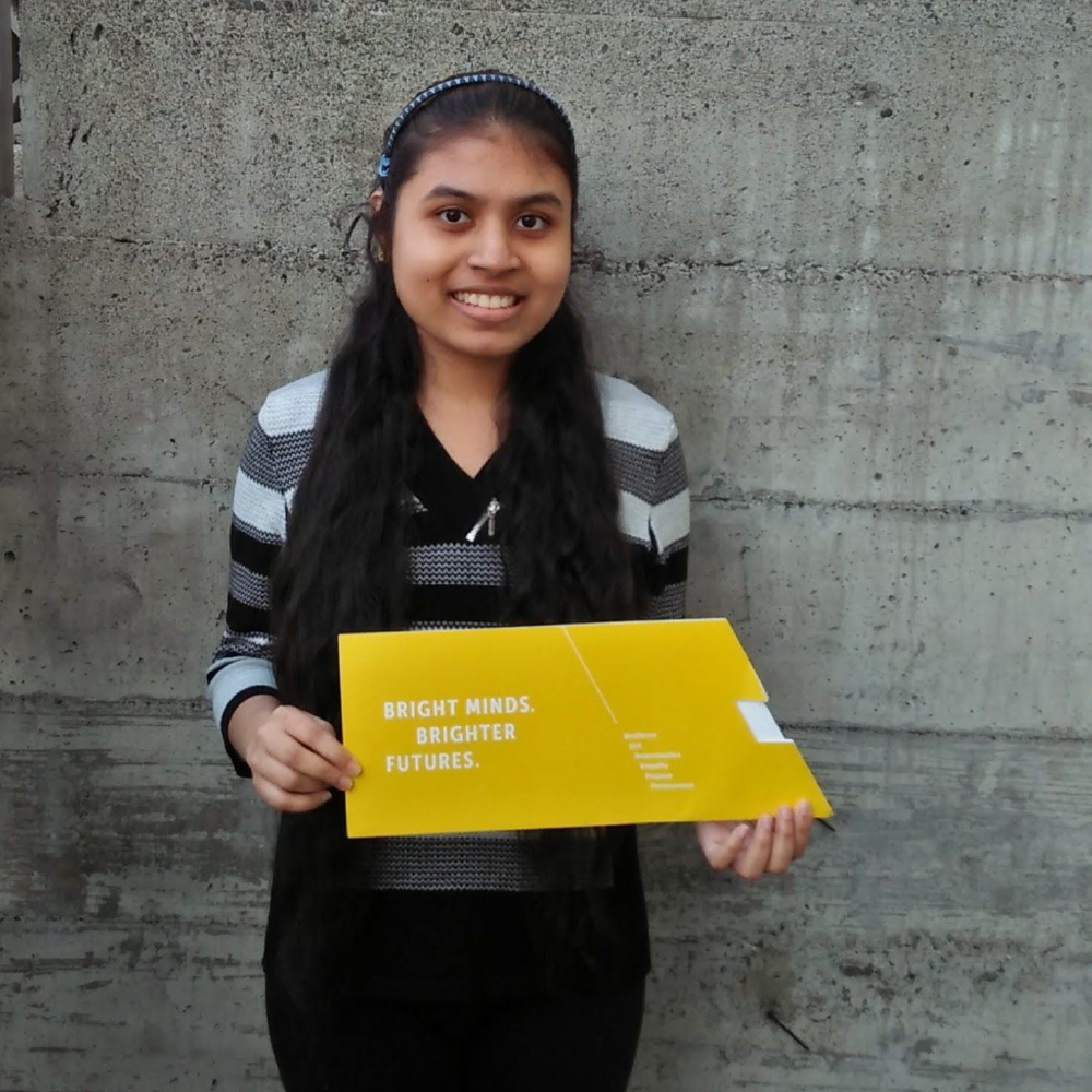 Woman stands against concrete background, smiling and holding yellow certificate proudly.