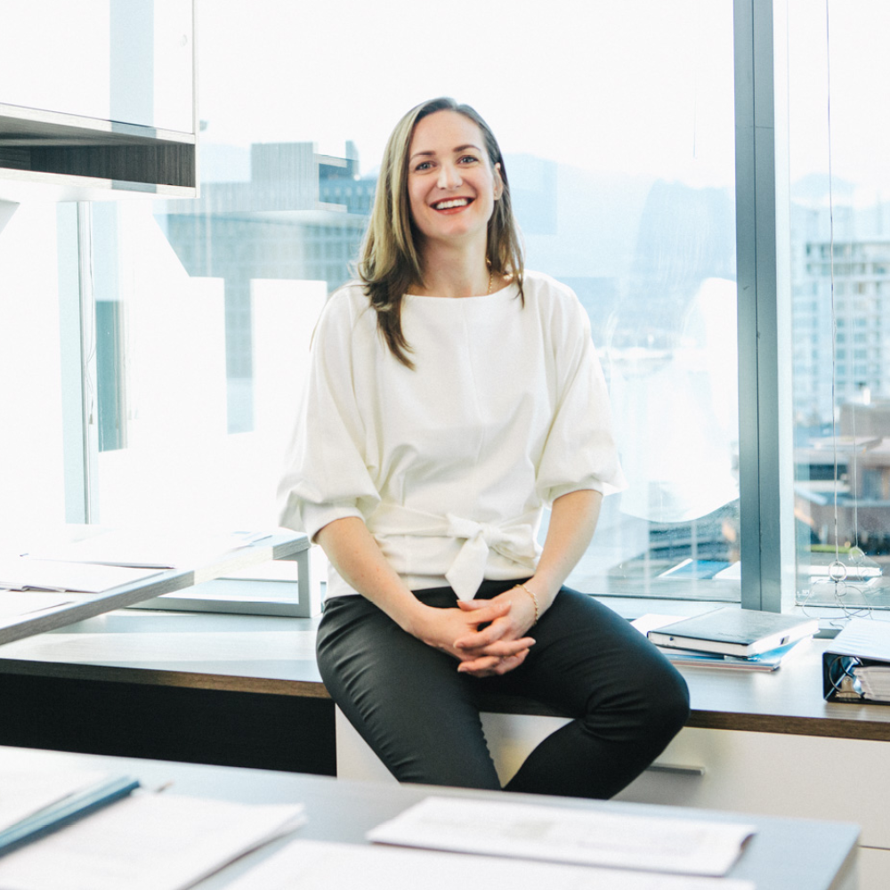 Woman in business casual wear sits on the ledge of a high-rise office space, smiling with hands joint together.