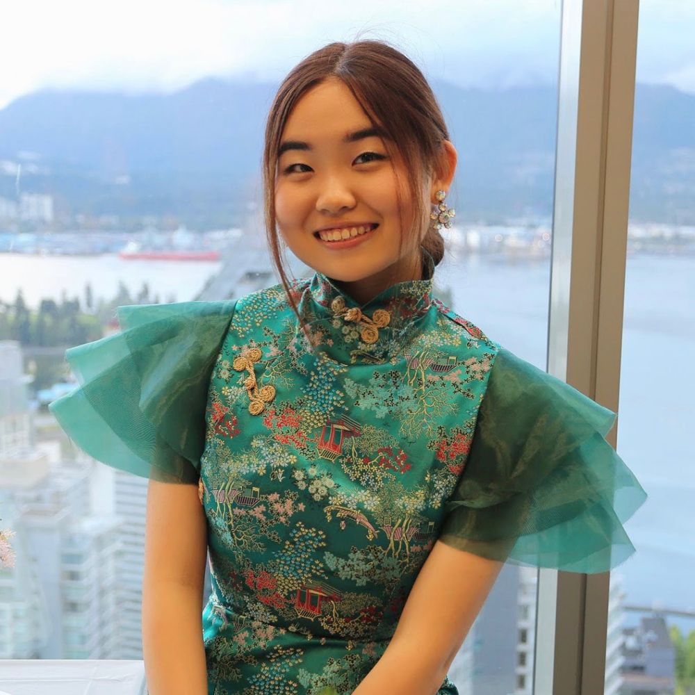 Woman wearing floral turqouise dress stands against a window, looking out to the water and mountains.