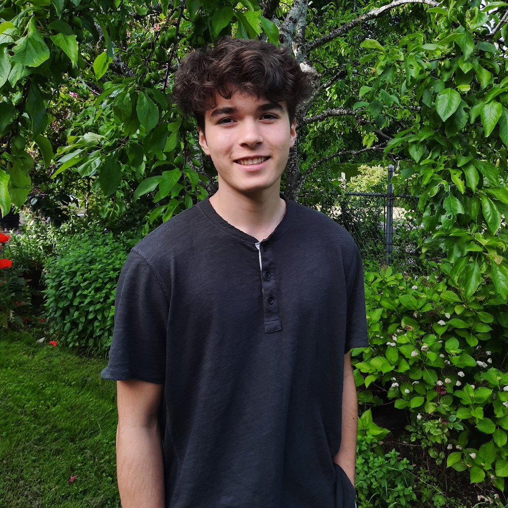 Young man with dark curly hair wears dark blue t-shirt, standing in front of greenery.