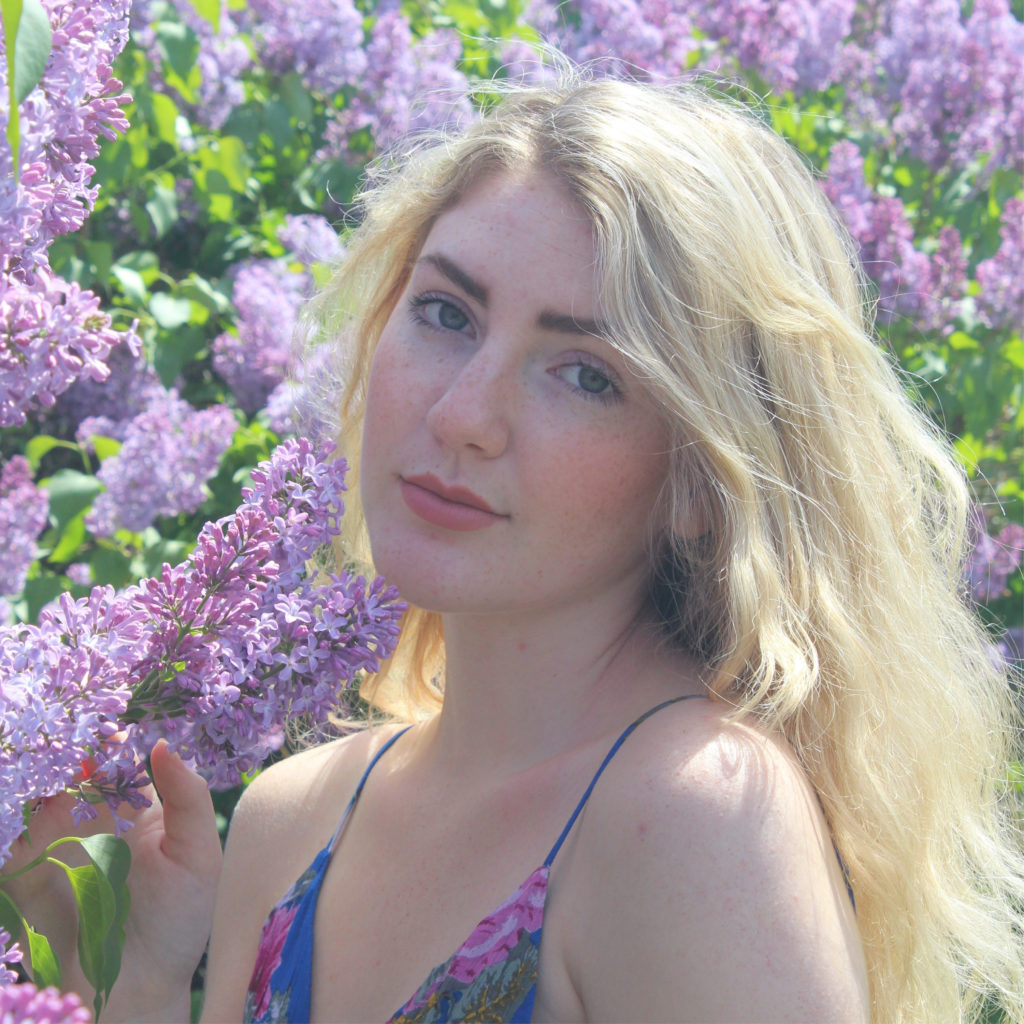 Young woman with blonde hair stands against purple flowers.
