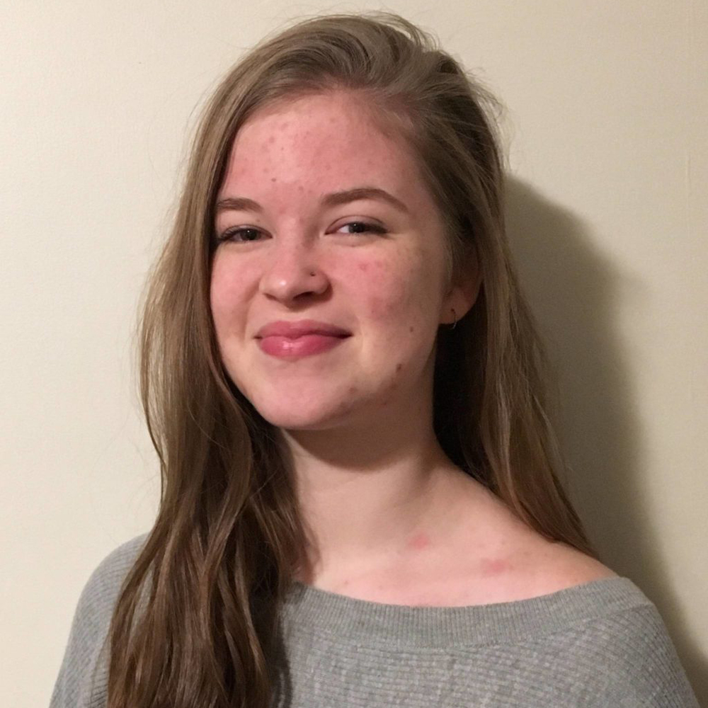 Young woman stands against a white wall, smiling at camera.