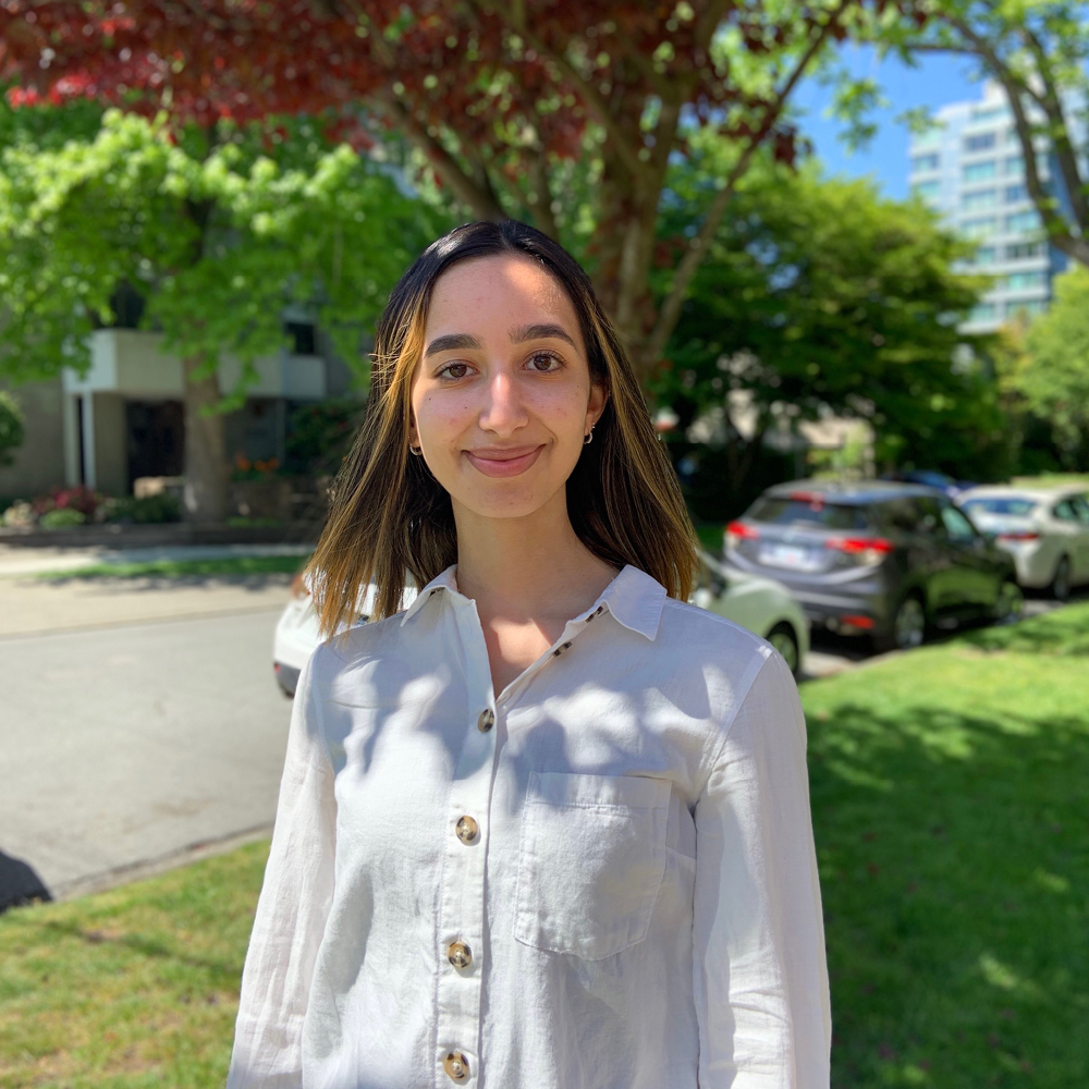 Young woman wearing white shirt stands on neighbourhood street, shaded by tree.