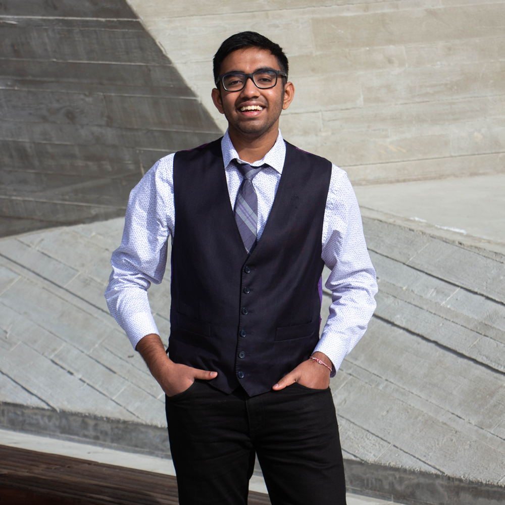 Young man wearing a dress shirt, tie, and vest, stands against a concrete background with hands in his pocekts.