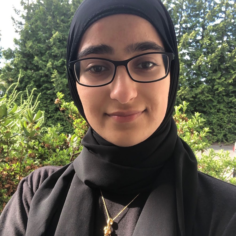 Woman wearing glasses and a black hijab smiles in a selfie.