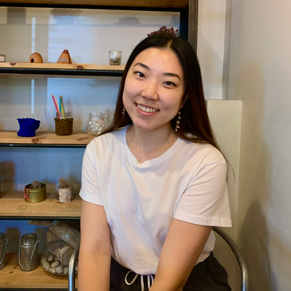 Young Asian woman sitting in a chair, wearing white t-shirt against a shelf at a cafe.