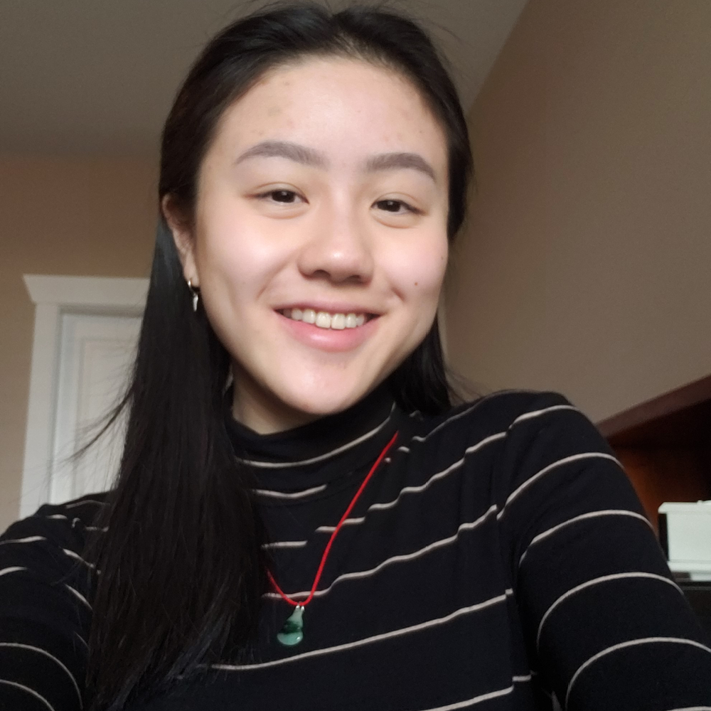 Young Asian woman in black t-shirt with white stripes, smiles in a selfie taken in a room.