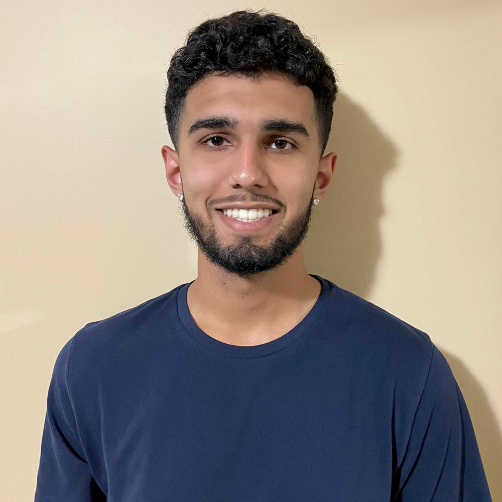 Young man with facial hair stands against a blank background, smiling with teeth in a navy shirt.
