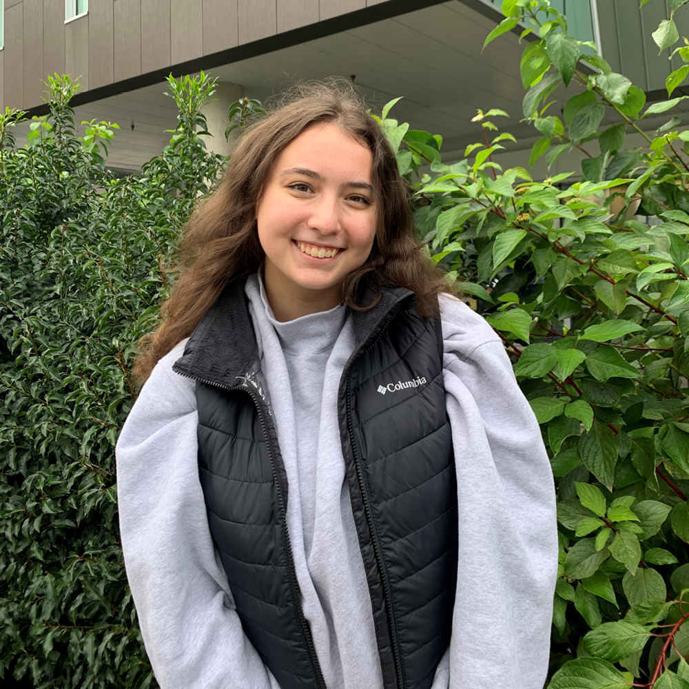Student wearing grey sweatshirt and black vest stands outside against greenery, smiling at the camera with her hands together.