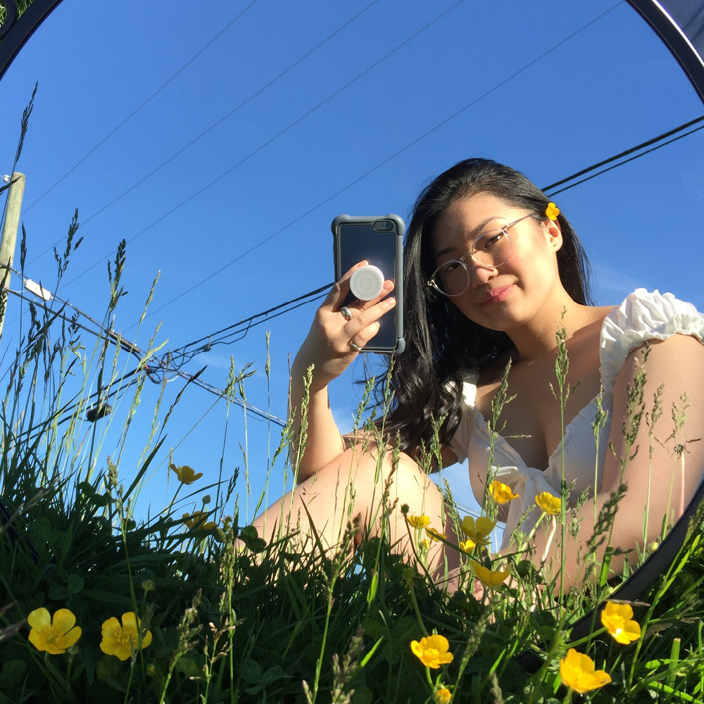 Young woman takes a mirror selfie in a grass field with yellow buttercup flowers.
