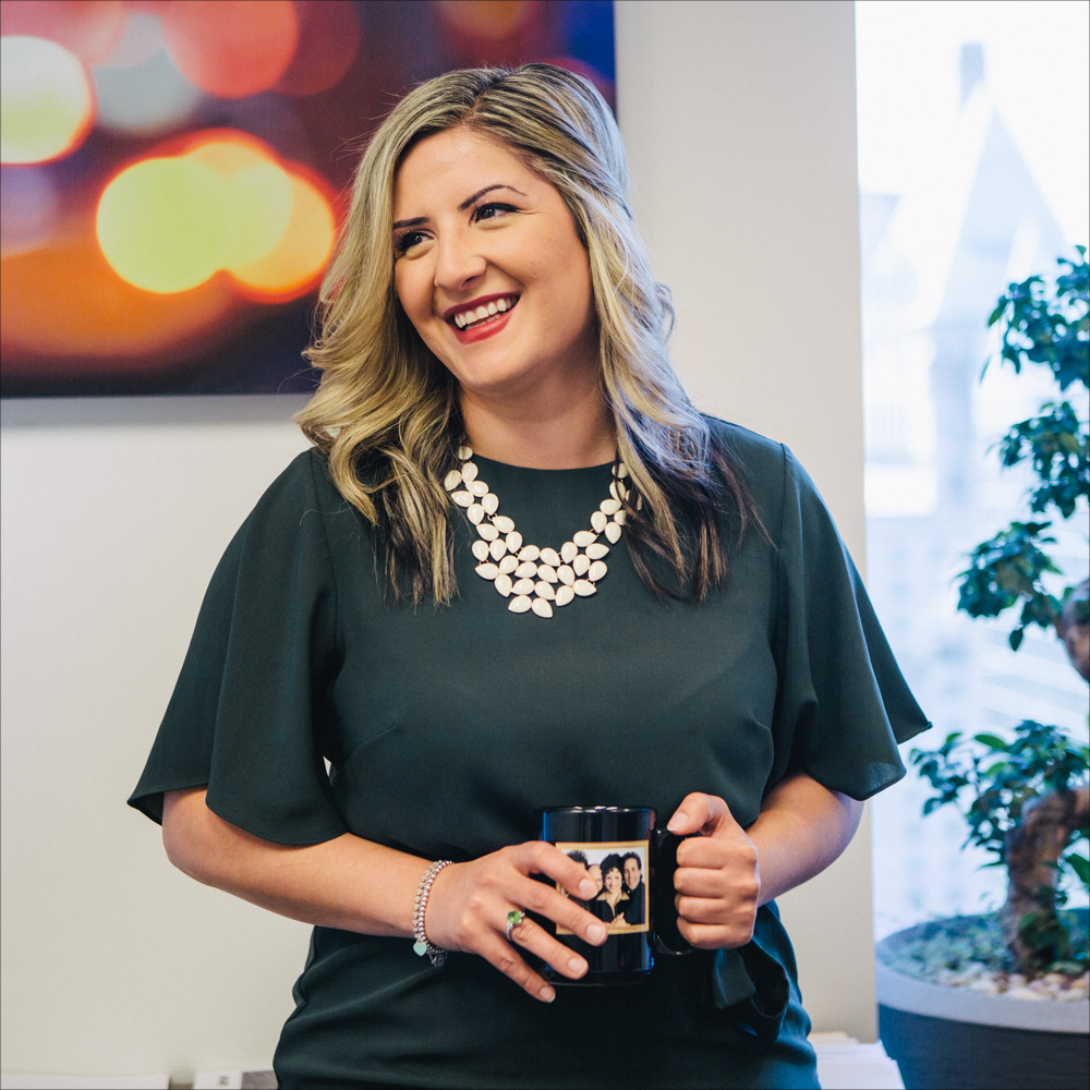 Woman in dark top and statement necklace stands while facing her right and smiling.