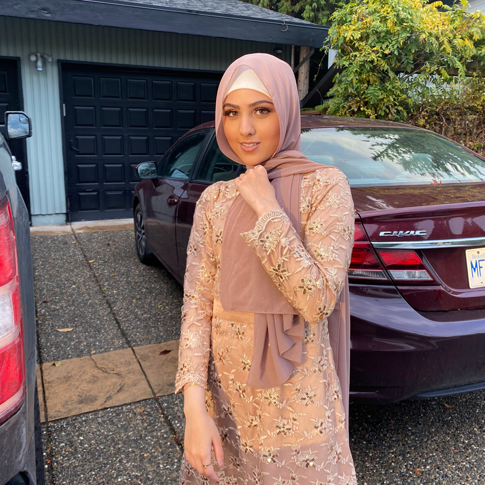 Woman in pink hijab stands in front of a burgundy sedan outside a house.