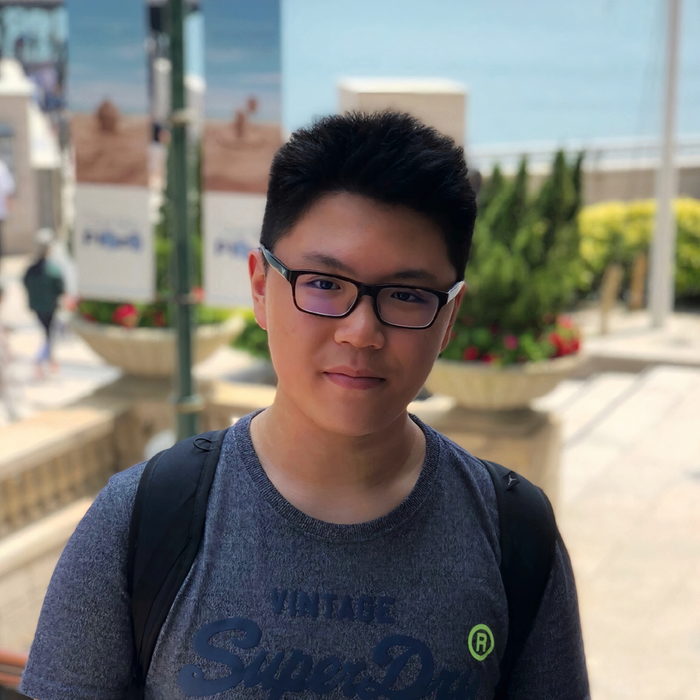 Young Asian man with black-frame glasses stands facing the camera, wearing a backpack.