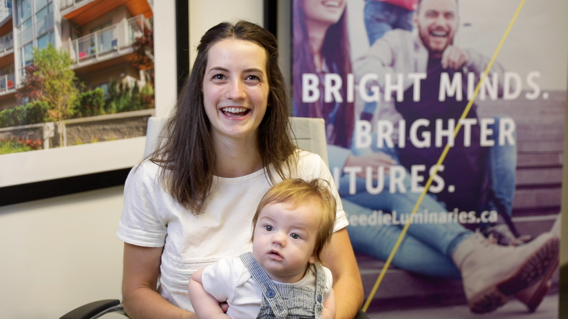 A young mother sits in front of a Beedie Luminaries banner, holding a toddler on her lap.