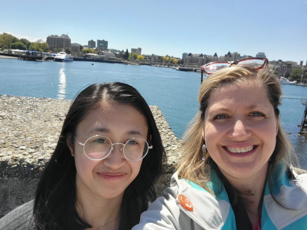 Student (left) and mentor (right) stand side-by-side, smiling in a selfie with a bright blue body of water in the background.