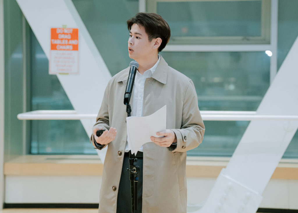 Male student presents a speech in front of a microphone, wearing a beige overcoat.