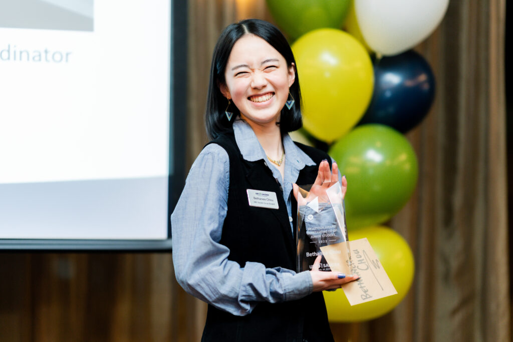 Bethanea, a young Asian woman, wearing a blue blouse and black vest, smiles proudly while holding a clear glass trophy. Behind her are balloons of various colours, representing Sauder School of Business at UBC.