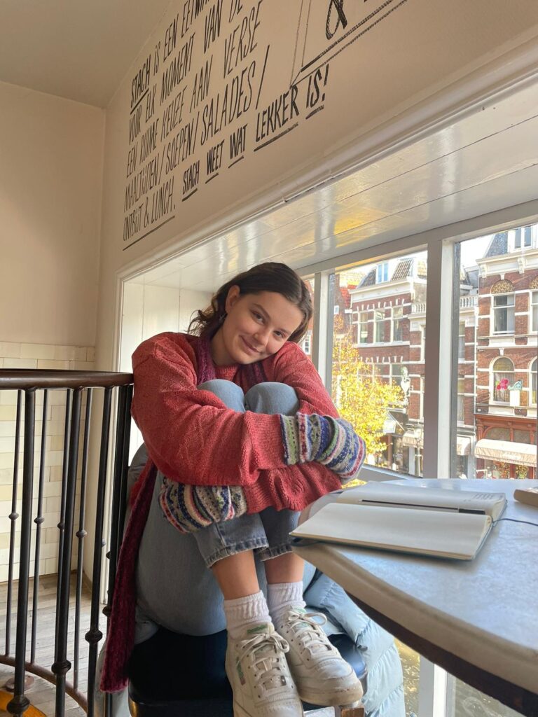 Tessa Brown Jones sits in a cafe on a sunny day, wearing a pink sweater and jeans. She hugs her knees, with her feet on the chair.