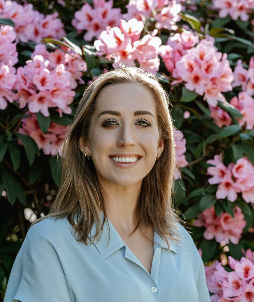 Headshot of Nicole Bailey, wearing a blue shirt and standing in front of blooming pink flowers.