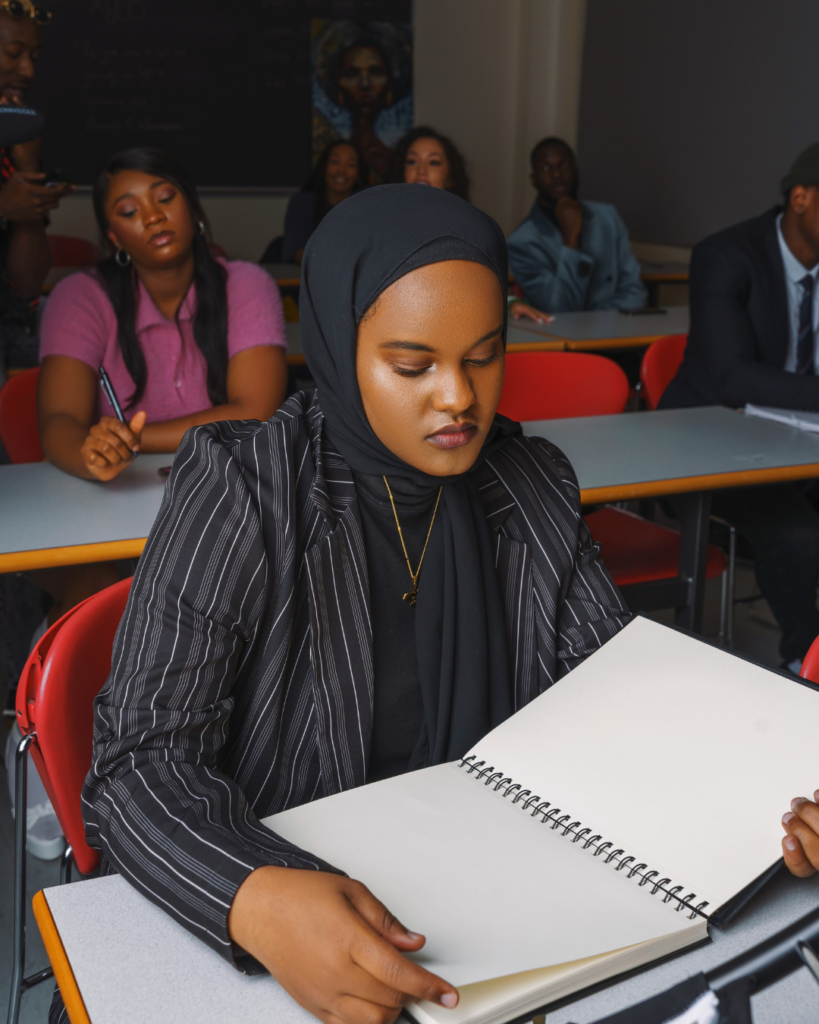 Female student in black hijab and blazer, looking down at a notebook in a classroom.