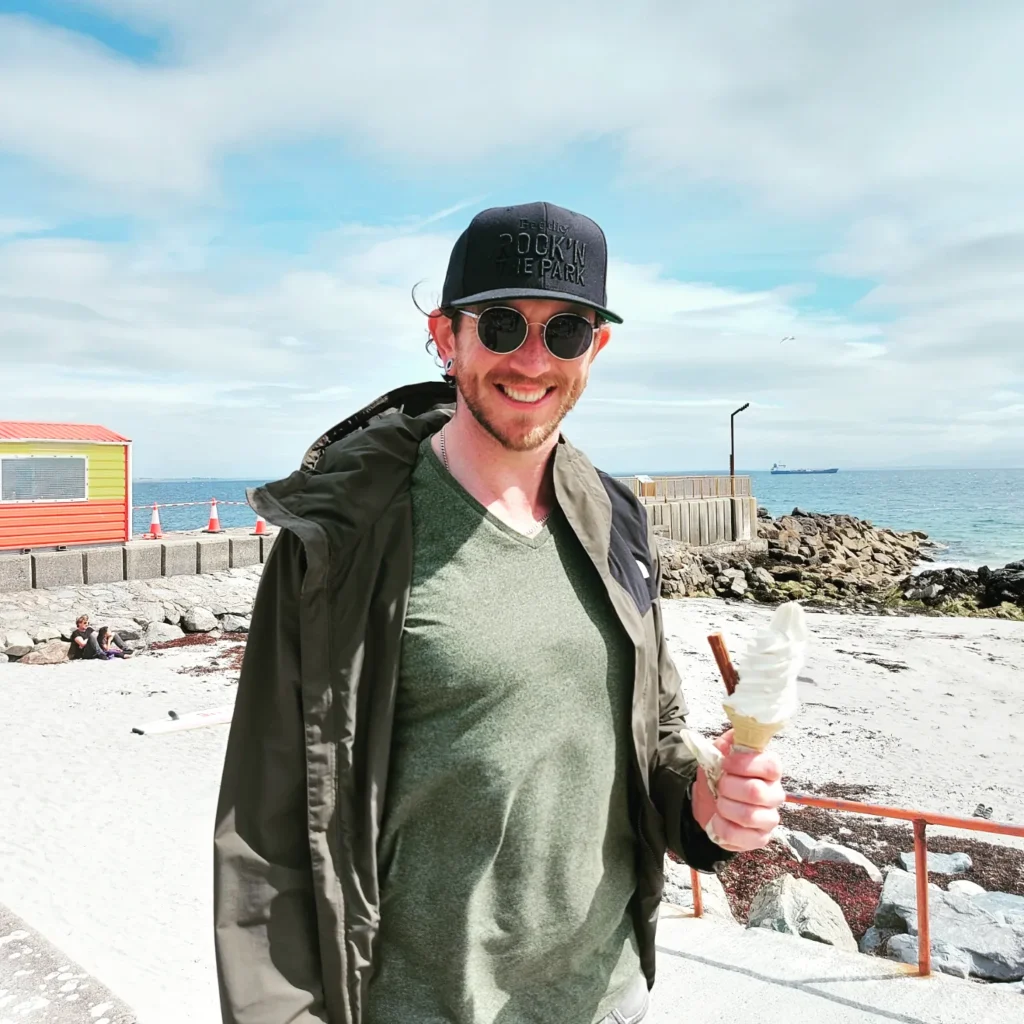 Jamie, wearing a jacket over his green shirt, holds on ice cream cone near the waterfront on a sunny day.