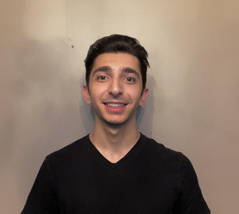 Young man stands against a blank wall, smiling.
