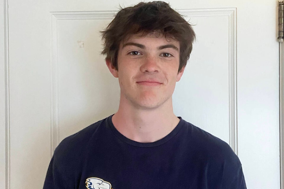 A young man stands against a door, smiling and wearing a black tee shirt.