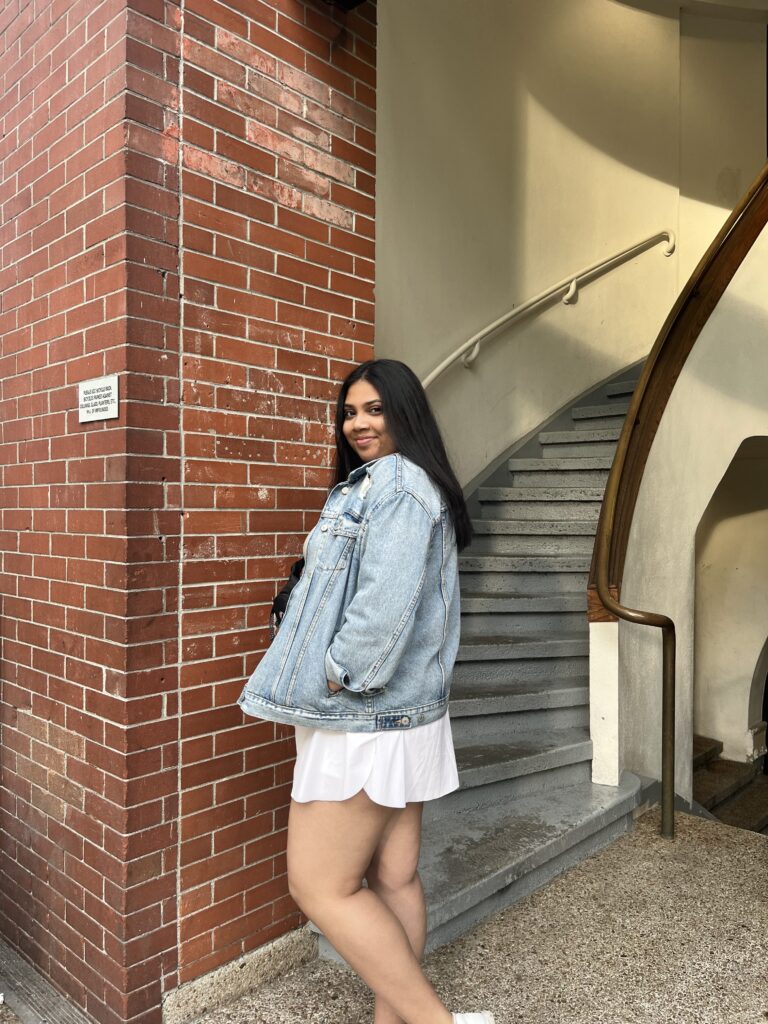 Srusti, wearing a white dress and denim jacket, poses against a staircase and red brick wall, looking back at the camera.