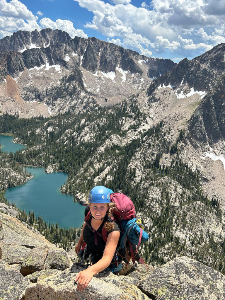 Avie wears a blue helmet and large backpack, while adventuring the backcountry terrain. Behind her are clouds, a large mountain range, and alpine lakes.