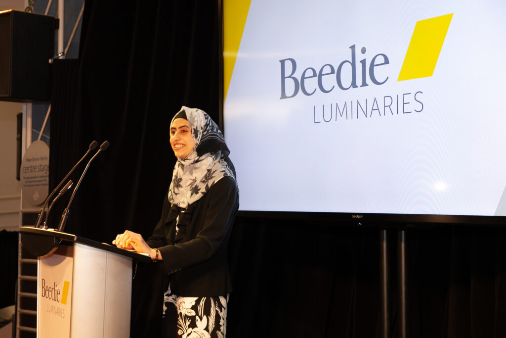 Woman in patterned hijab and black clothing stands at a podium, presenting in front of a Beedie Luminaries slide show.