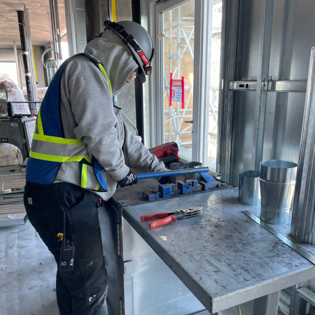 Ashleigh Roy in a safety vest and goggles, working on sheet metal on a construction site.