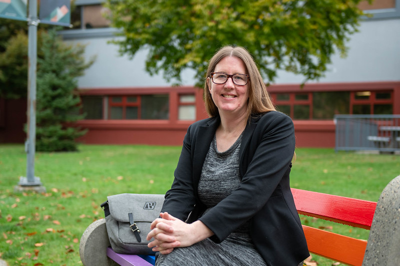 Gina sits on a red bench, smiling and wearing a black blazer. To her left is the Beedie Luminaries laptop bag.