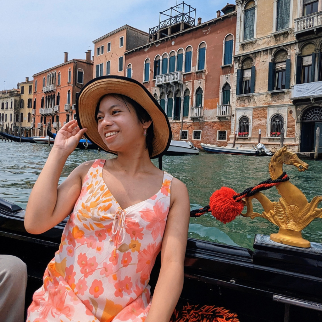 Diane sits on a boat, with European-style buildings behind her, wearing a floral orange dress.