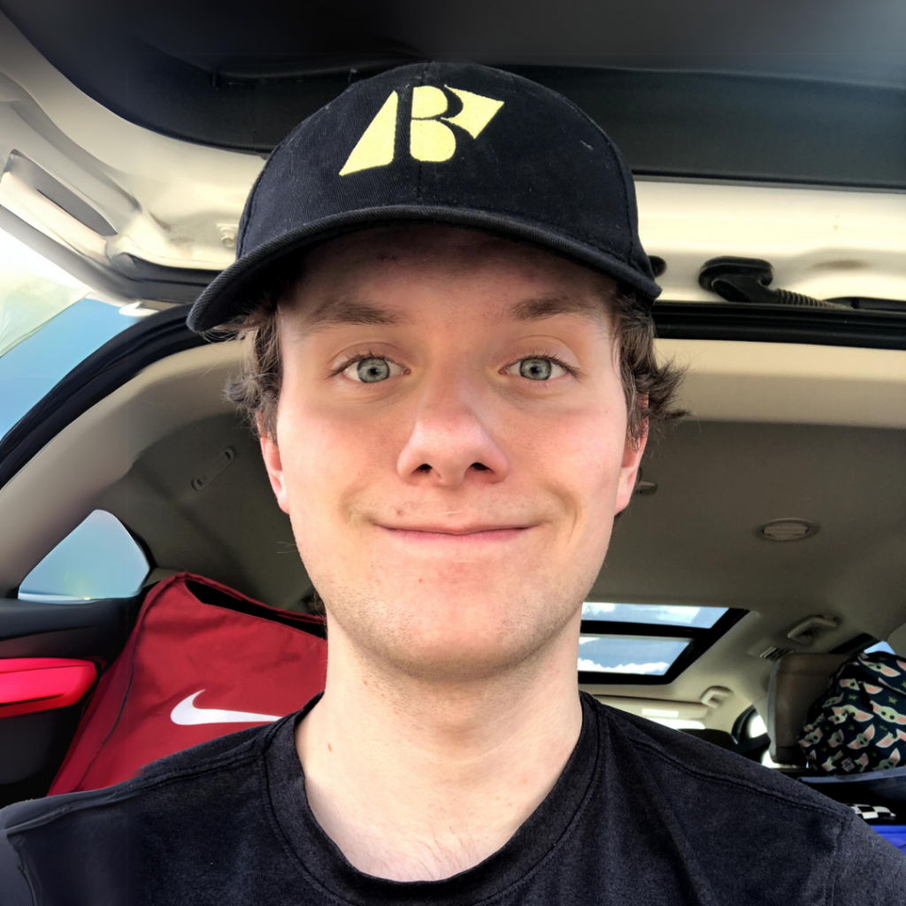 Cain smiles in a selfie taken in his car, wearing a black hat featuring the yellow Beedie Luminaries logo.