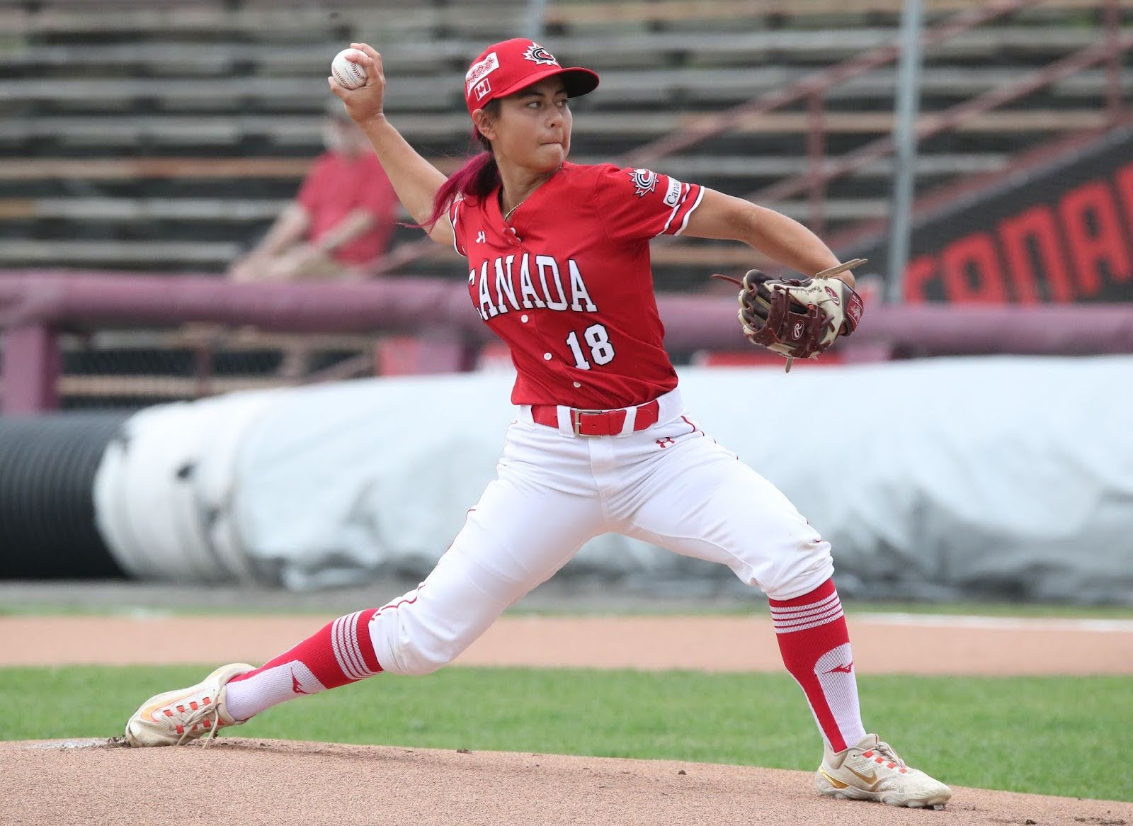 Raine, wearing team uniform of white pants and a bright red jersey, is pictured pitching the ball on a baseball field.