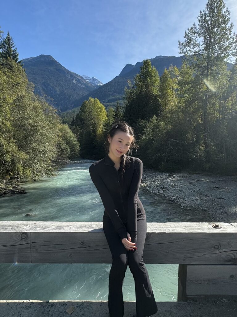 Tina Zeng, wearing all black, sits on a wooden bridge with a river, trees, and mountains behind her on a sunny day.